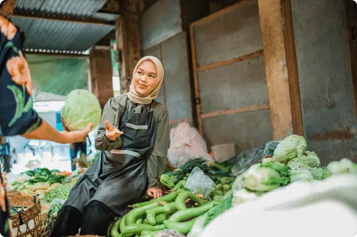 A market trader in a headscarf offering a cabbage to a customer at her vegetable stall